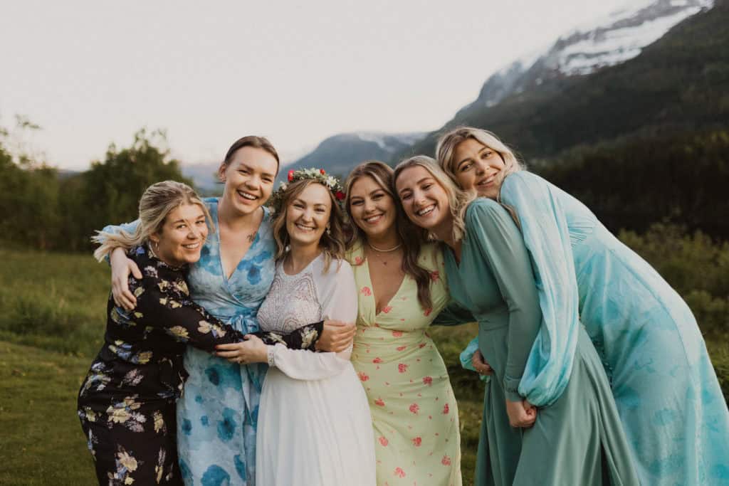 kvinner i fargerike kjoler og et i hvit kjole klemmer, med fjell og natur bak dem