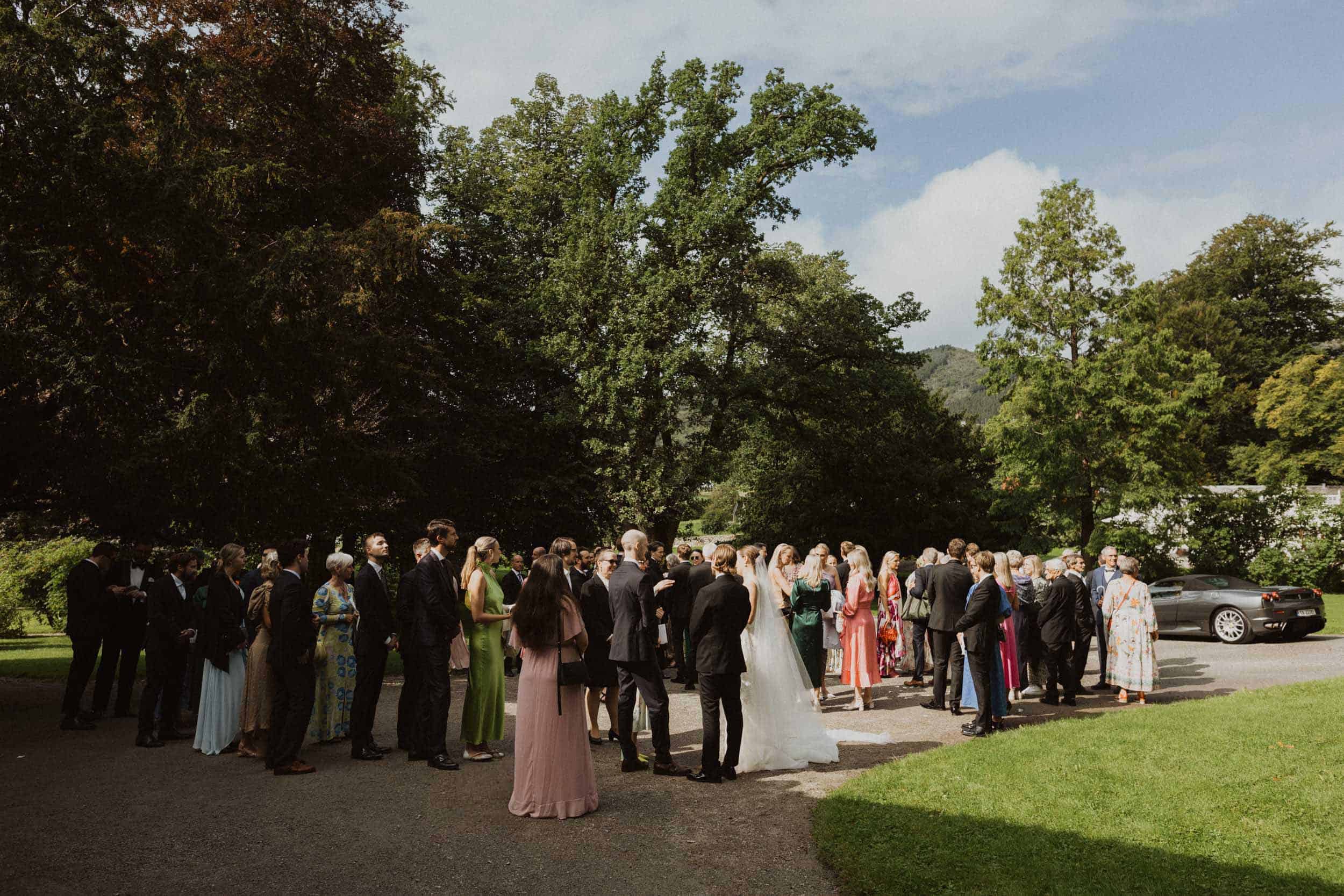 mange mennesker i fine klær i en park og et brudepar i midten