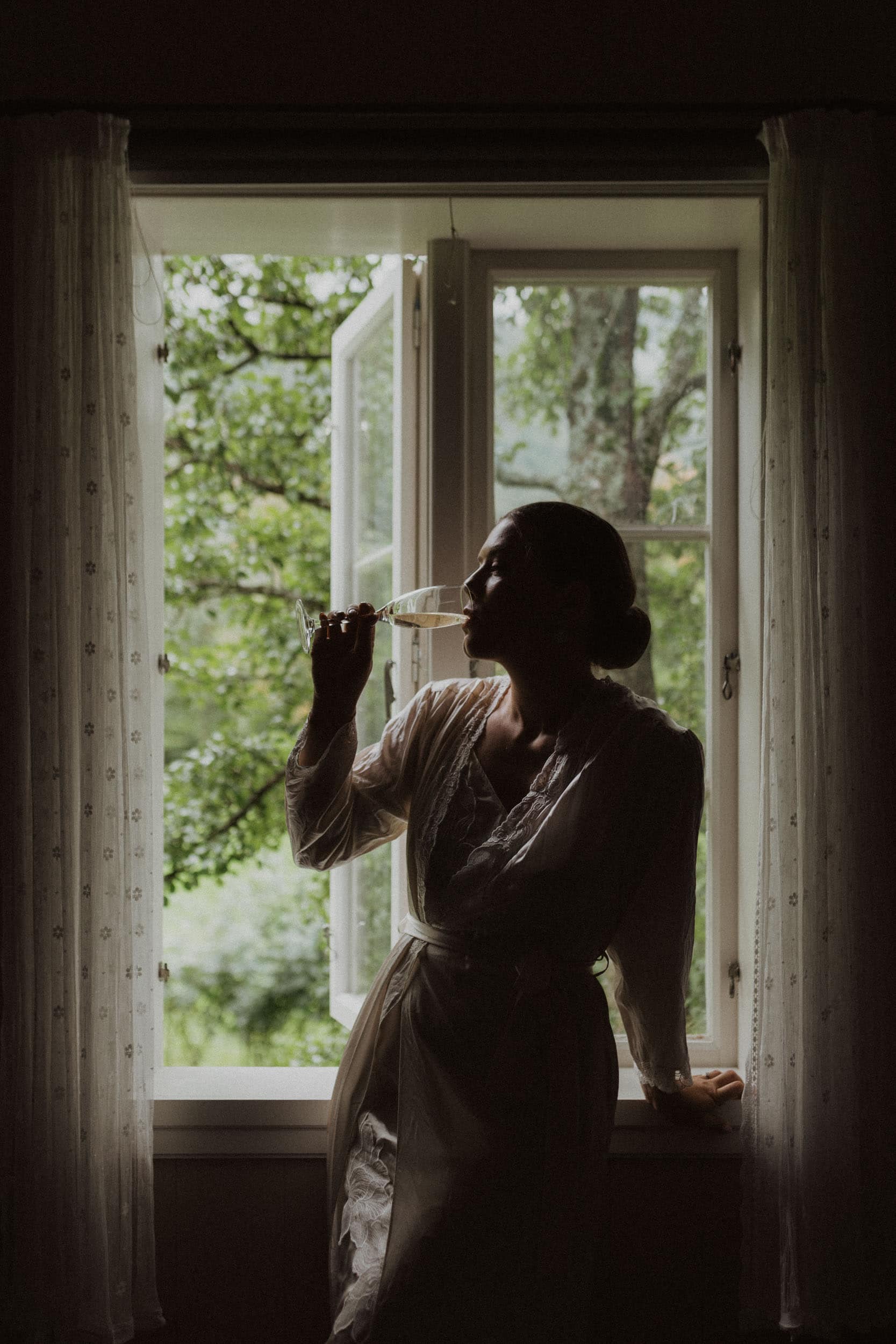 kvinne som drikker et glass champagne sitter i vinduskarmen og grønne trær bak vinduet