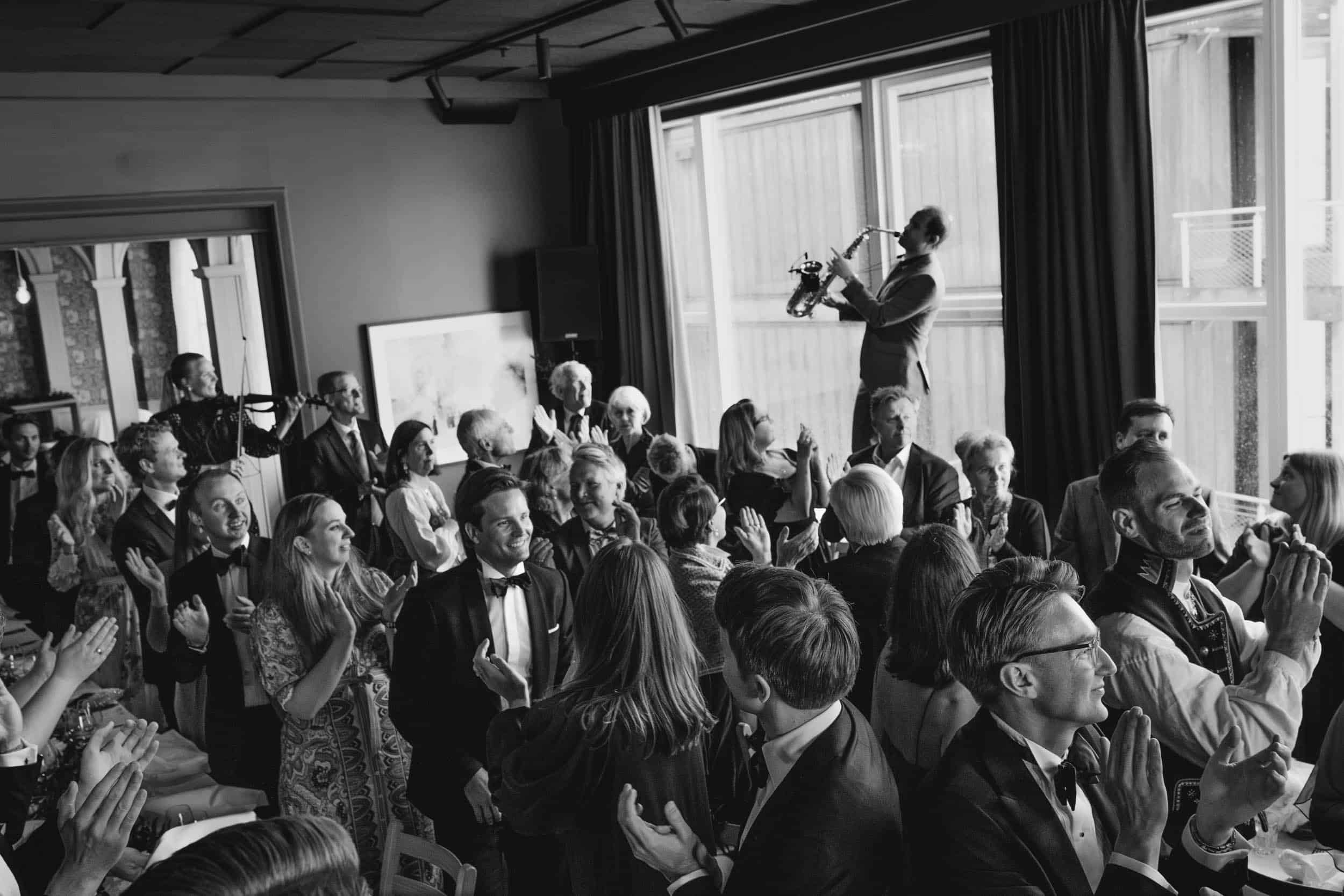 mange mennesker i fine klær synger og klapper til saksofonist som spiller for dem